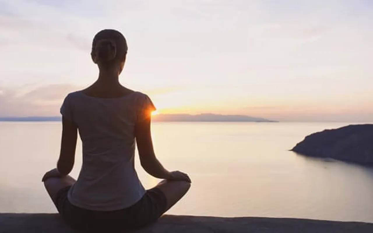 woman meditating while sitting in front of water in a lotus position with the sunset at the far background